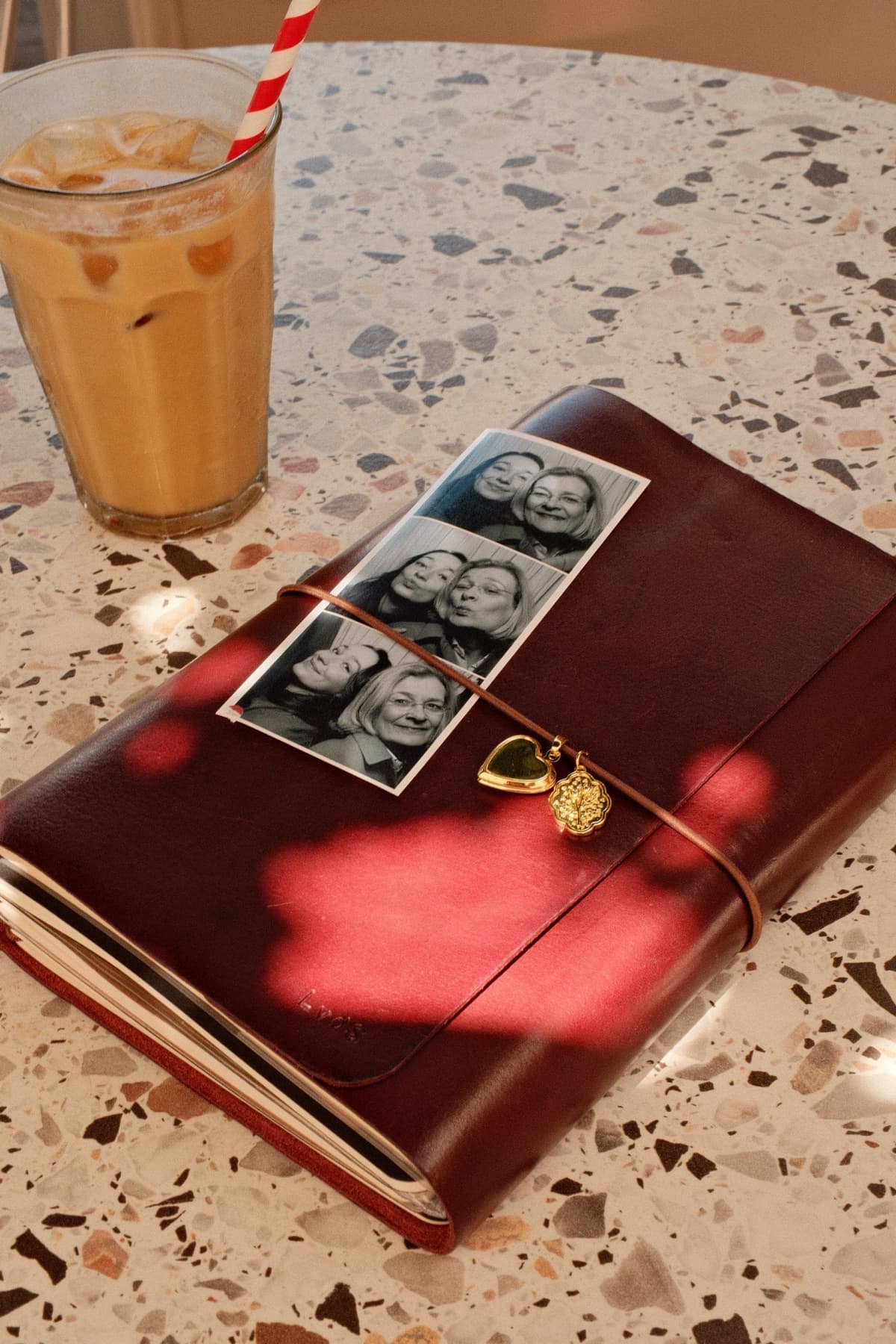 Een wijnrode Club YUP-journal met fotostrip en gouden bedels op een terrazzotafel naast een ijskoffie